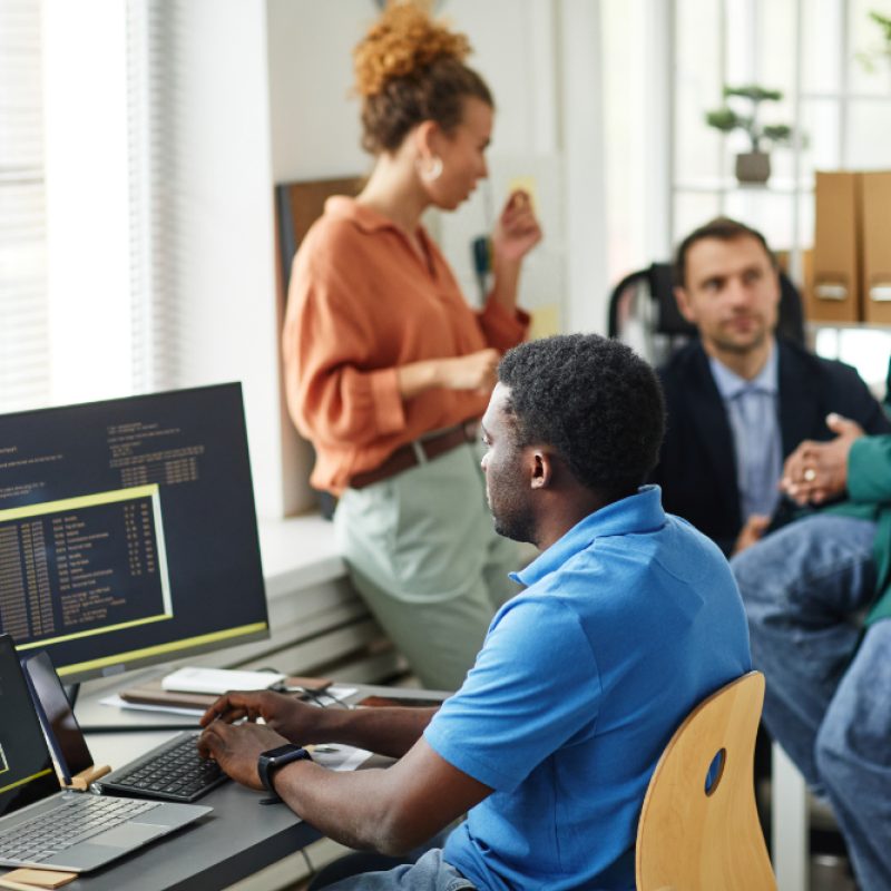 African IT specialist working with software typing on keyboard sitting at table with computers while his colleagues working in team in background